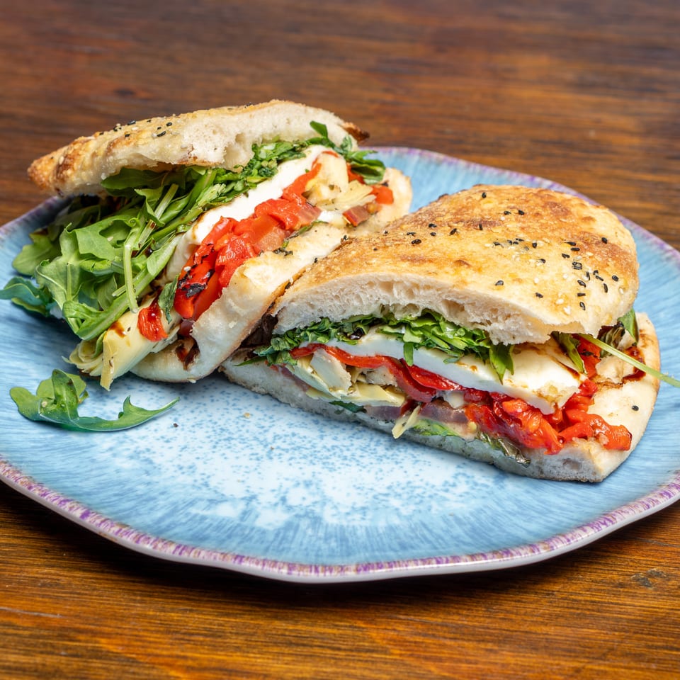 Two halves of a focaccia sandwich filled with arugula, roasted red peppers, and cheese on a blue plate.