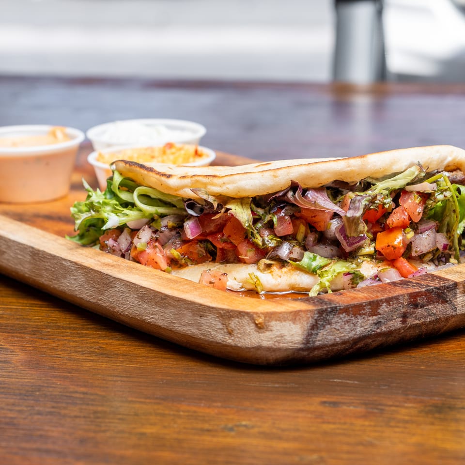 A pita bread sandwich filled with lettuce, tomatoes, and onions, served with two dipping sauces.