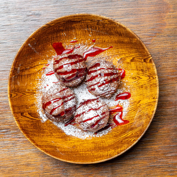 Four chocolate cream puffs with raspberry drizzle and powdered sugar on a wooden plate.
