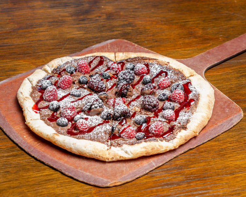 Dessert pizza with chocolate spread, raspberries, blueberries, blackberries, powdered sugar, and berry drizzle.