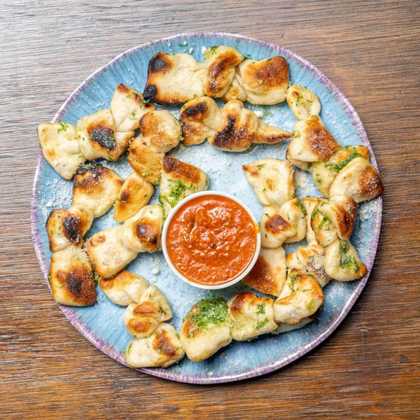 Garlic knots with parmesan and parsley, arranged around a small bowl of marinara sauce on a blue plate.