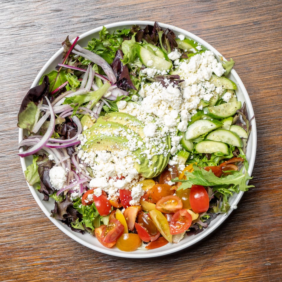 A vibrant salad with mixed greens, red onions, avocado, cucumbers, cherry tomatoes, and feta cheese.