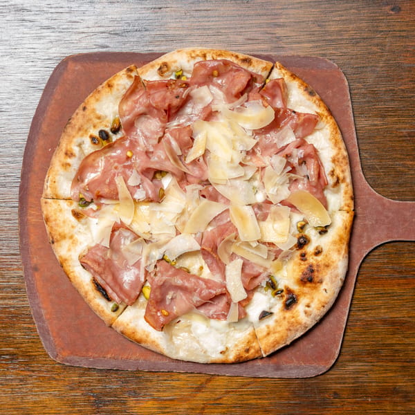 A whole pizza with prosciutto, shaved cheese, and pistachios on a wooden pizza peel.