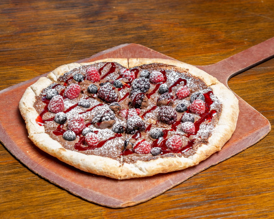 Dessert pizza with chocolate spread, raspberries, blueberries, blackberries, and powdered sugar on a wooden peel.